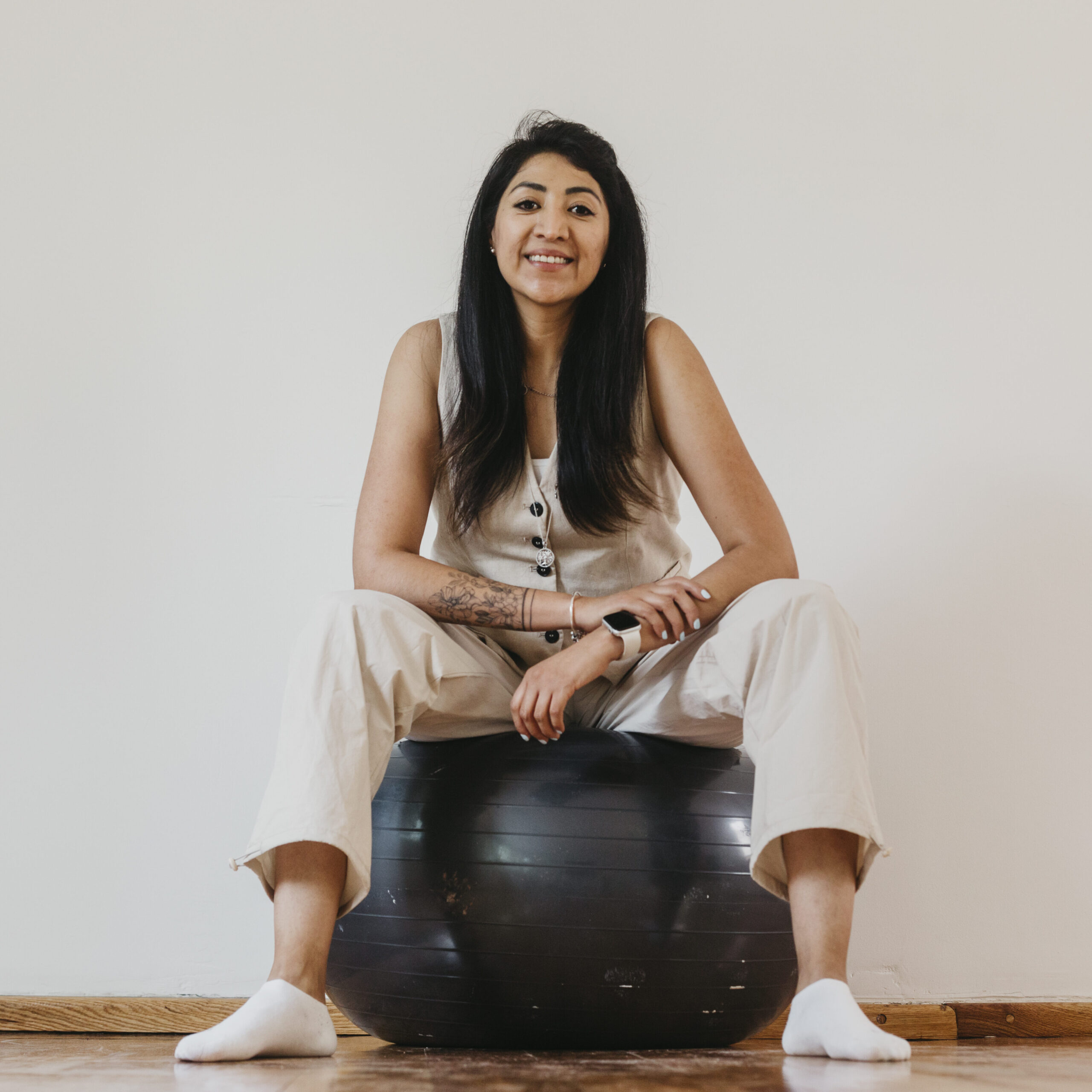 Mujer con ropa neutra sentada sobre una pelota de ejercicio, sonriente y en actitud cercana y empática